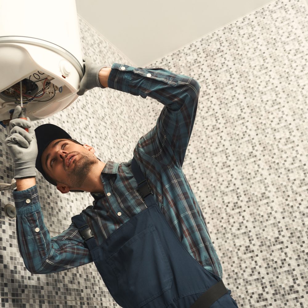 Plumber installing electric heating boiler. Young handyman in uniform setting up electric heating Plumber installing electric heating boiler. Young handyman in uniform setting up electric heating